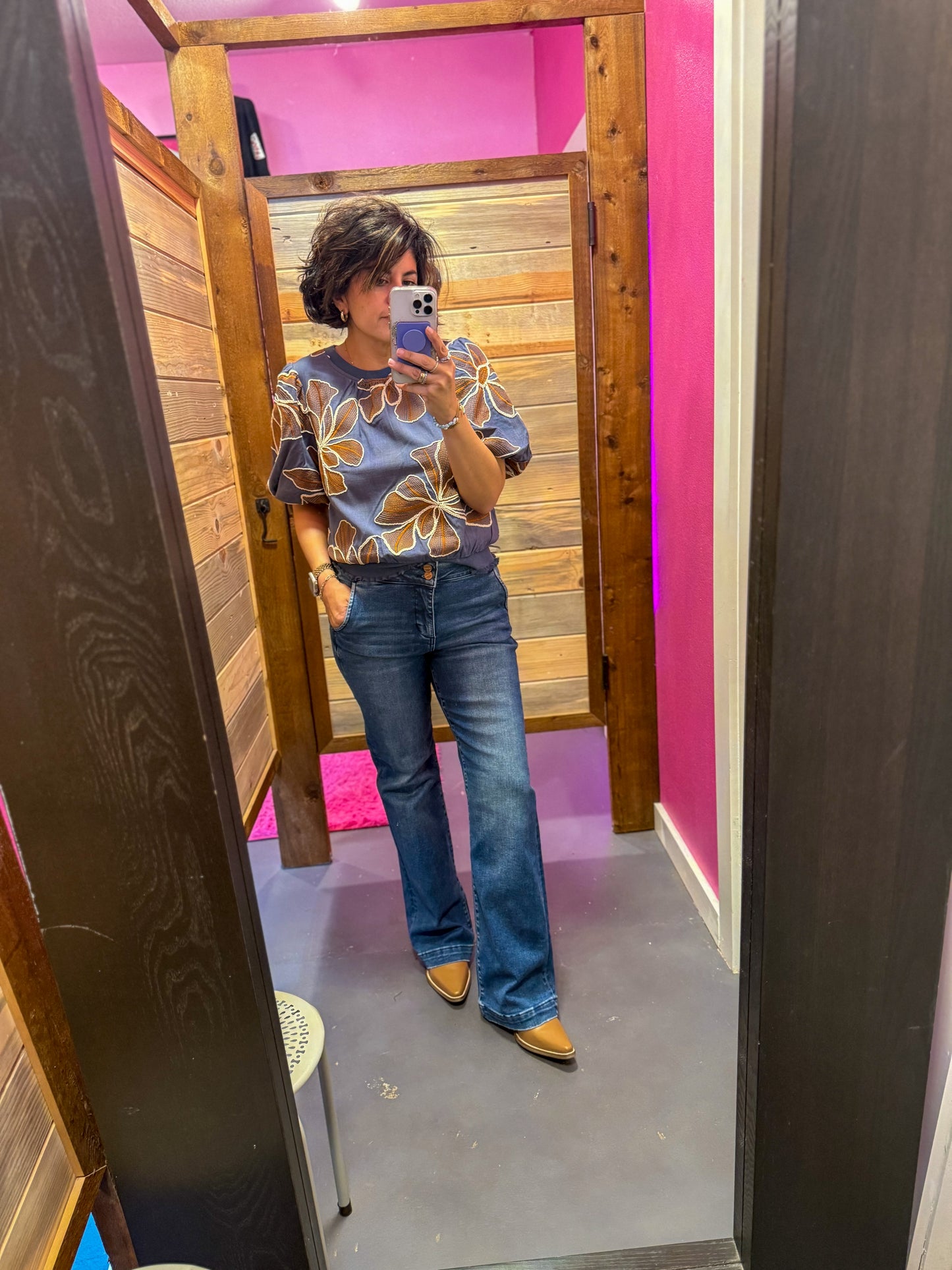Person taking a mirror selfie wearing a floral top and blue jeans in a room with pink walls.