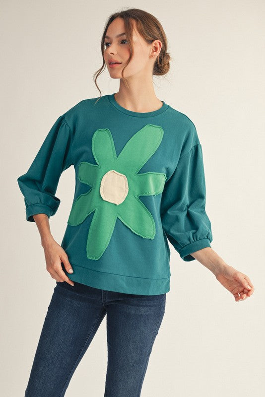 Woman wearing a teal sweatshirt with a large green flower design on a plain background