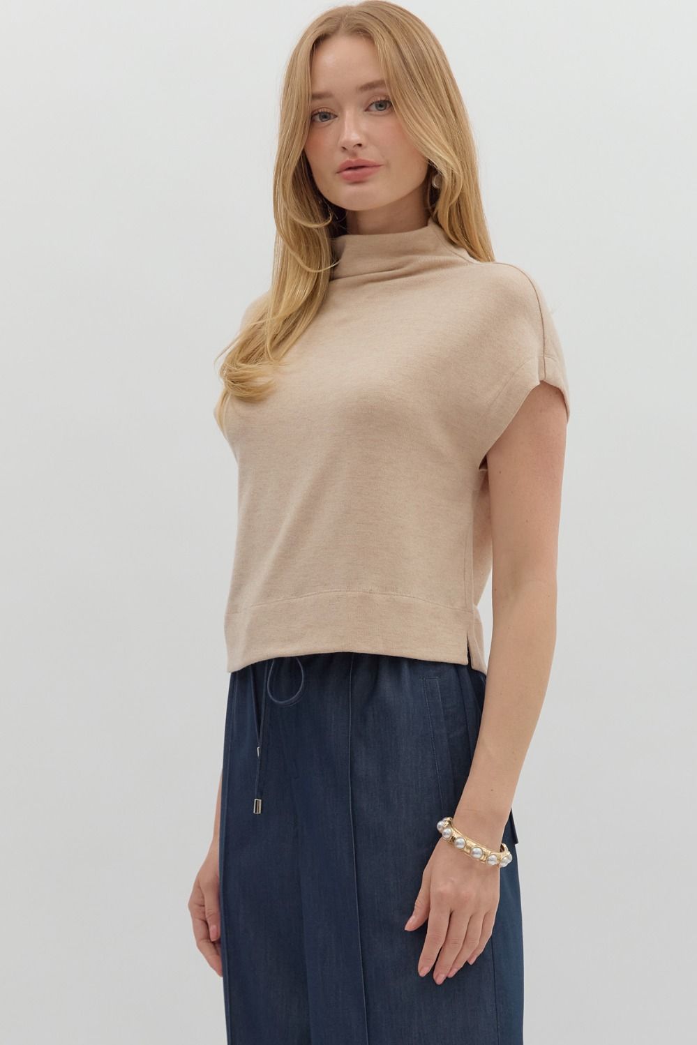 Woman wearing a beige turtleneck top and dark blue pants on a light gray background