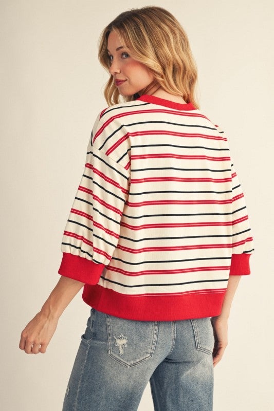 A woman wearing a red, white, and black striped knit sweater with red ribbed trim, paired with blue jeans, photographed against a plain light beige studio background.