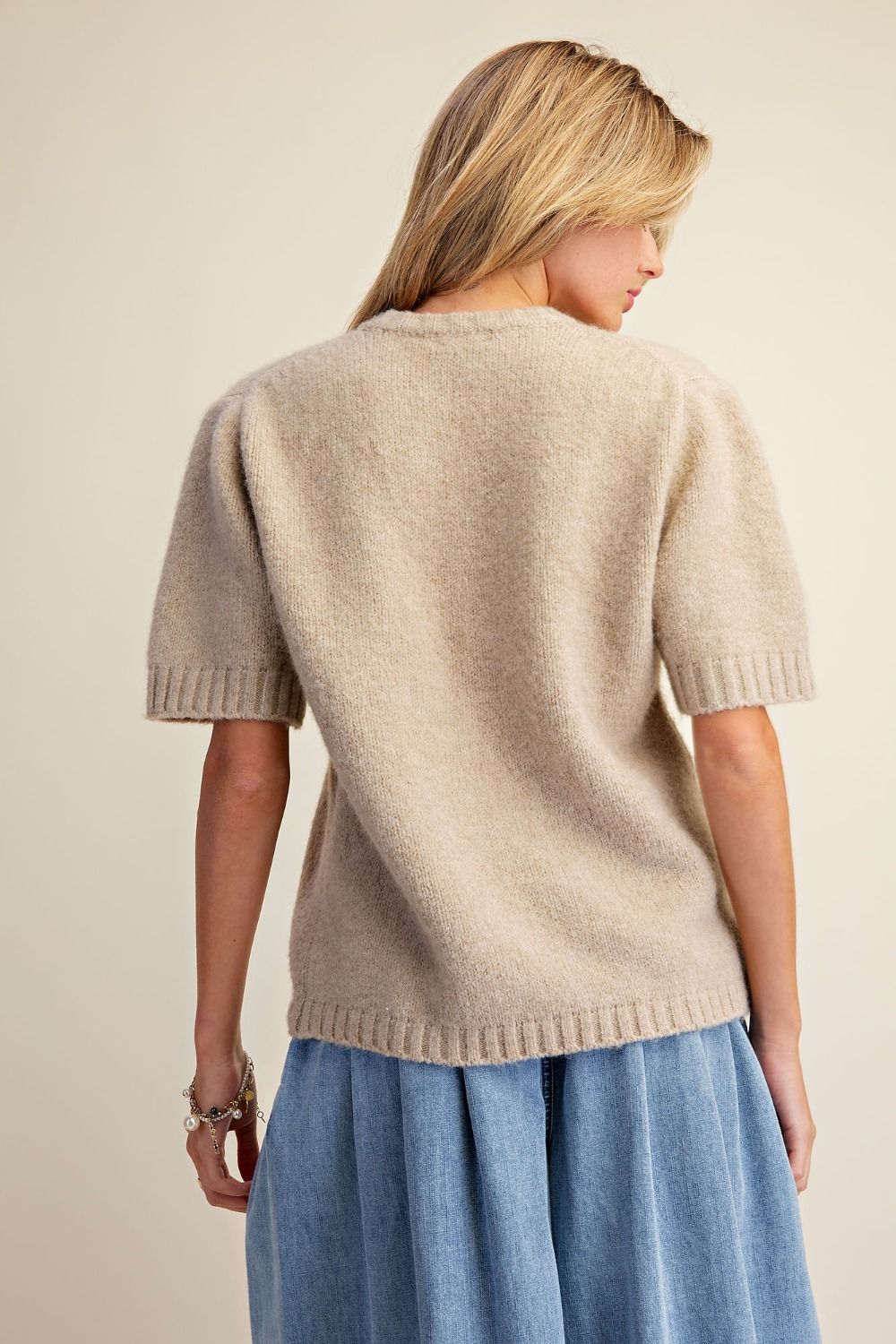 A woman shown from the back wearing a short-sleeve knit sweater in a light beige color, paired with a blue skirt, against a plain beige background.