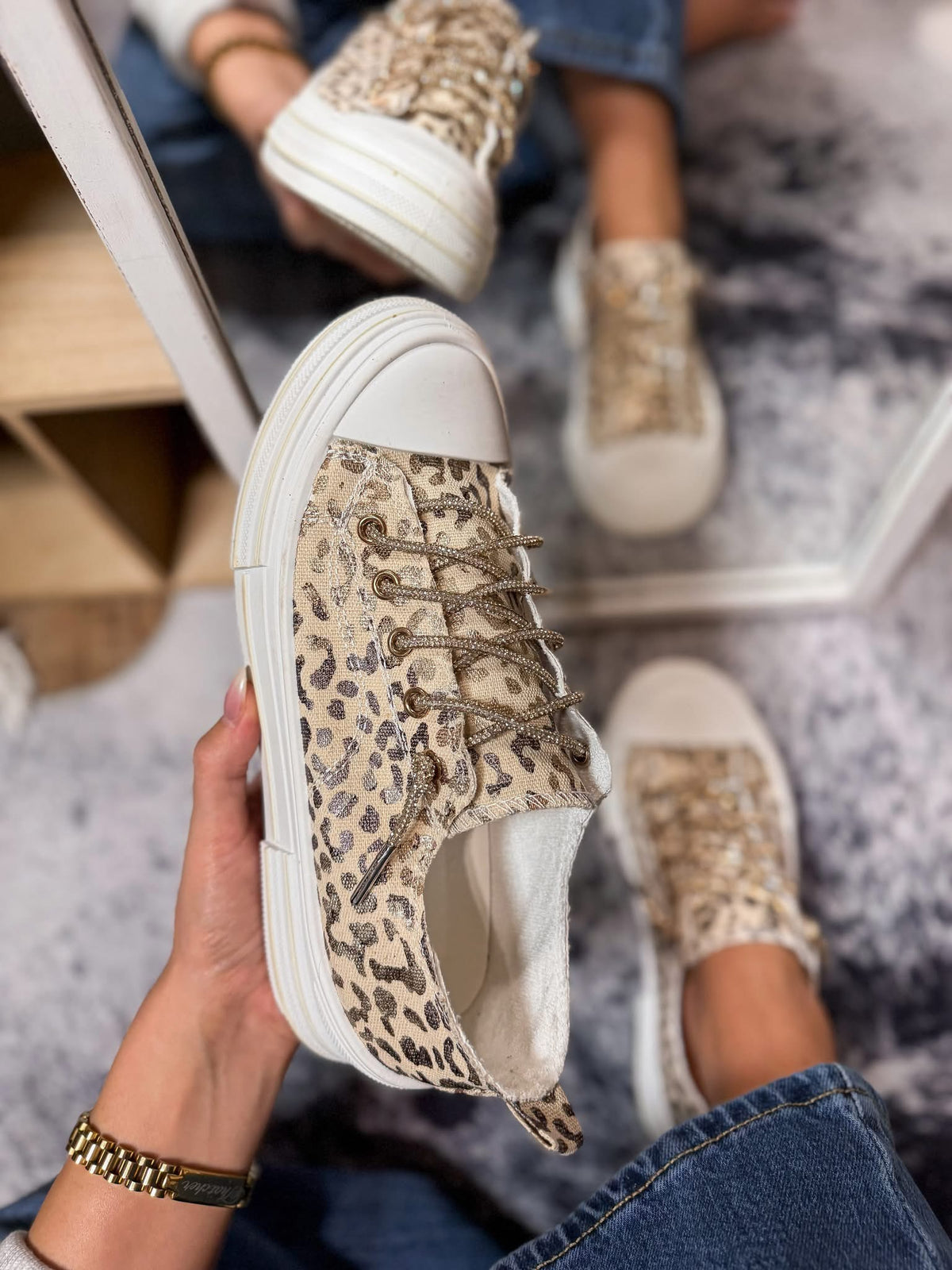 A person holds up a beige and brown leopard-print low-top sneaker with white soles and laces, while wearing the same sneakers with blue jeans in a mirror reflection.