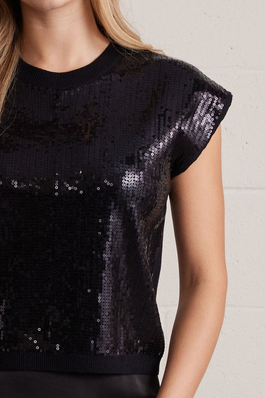A close-up of a woman wearing a black short-sleeve knit top covered in shimmering sequins, against a light neutral wall.