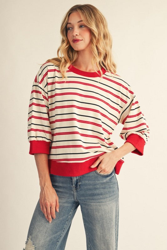 A woman modeling a red, black, and white striped crewneck sweatshirt with red ribbed trim, paired with blue jeans, against a plain beige background.
