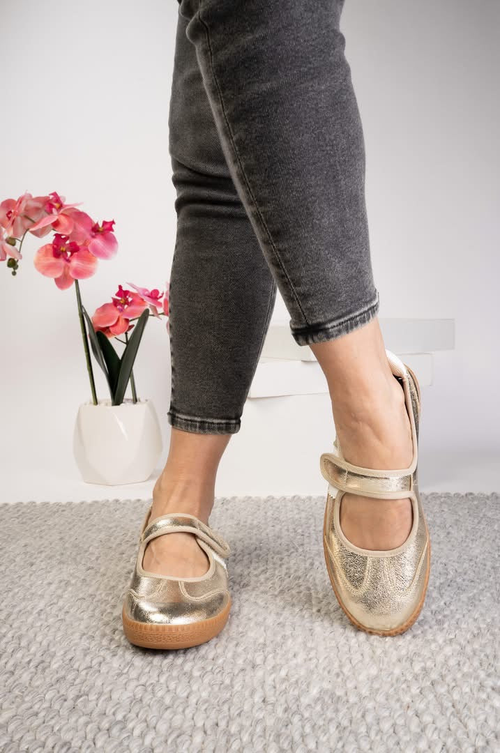 A person wearing metallic gold Mary Jane style shoes with dark gray cuffed jeans, standing on a light gray textured rug with a white vase of pink flowers in the background.