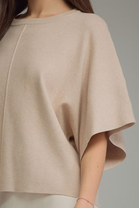 A close-up of a person wearing a light beige knit poncho-style top with wide draped sleeves and a crew neckline, shown against a plain gray background.