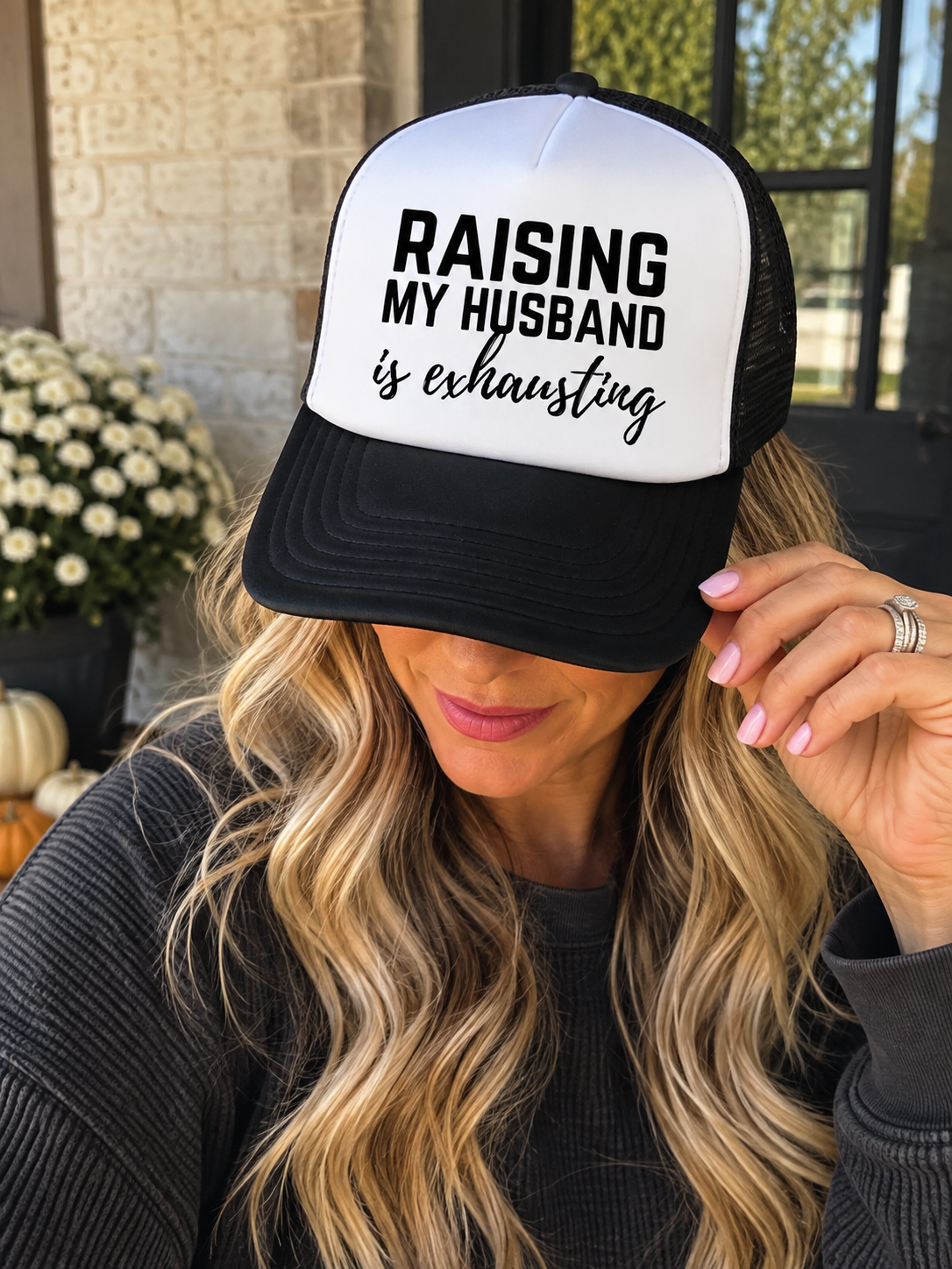Woman wearing a black and white Raising My Husband Is Exhausting trucker hat in a casual outdoor lifestyle setting.