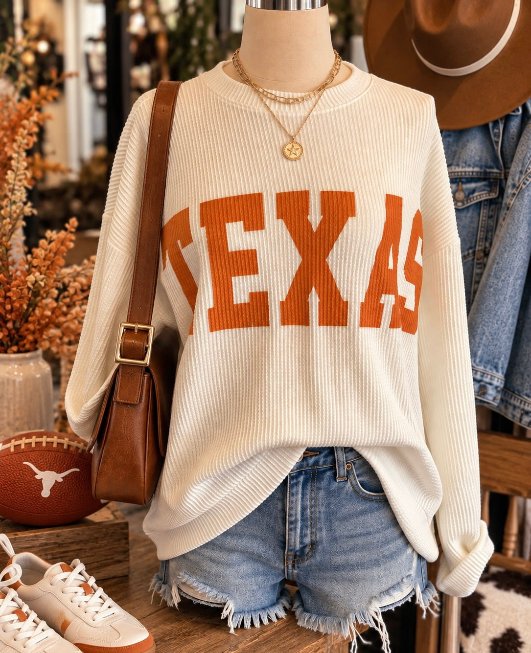 An off-white ribbed knit sweater with 'TEXAS' in orange varsity letters across the chest, styled on a mannequin with denim shorts, a brown shoulder bag, and layered gold necklaces in a boutique setting.