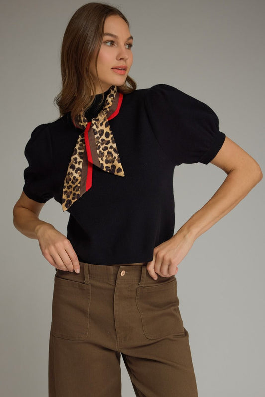 Woman wearing a black top with a leopard print scarf and brown pants on a gray background