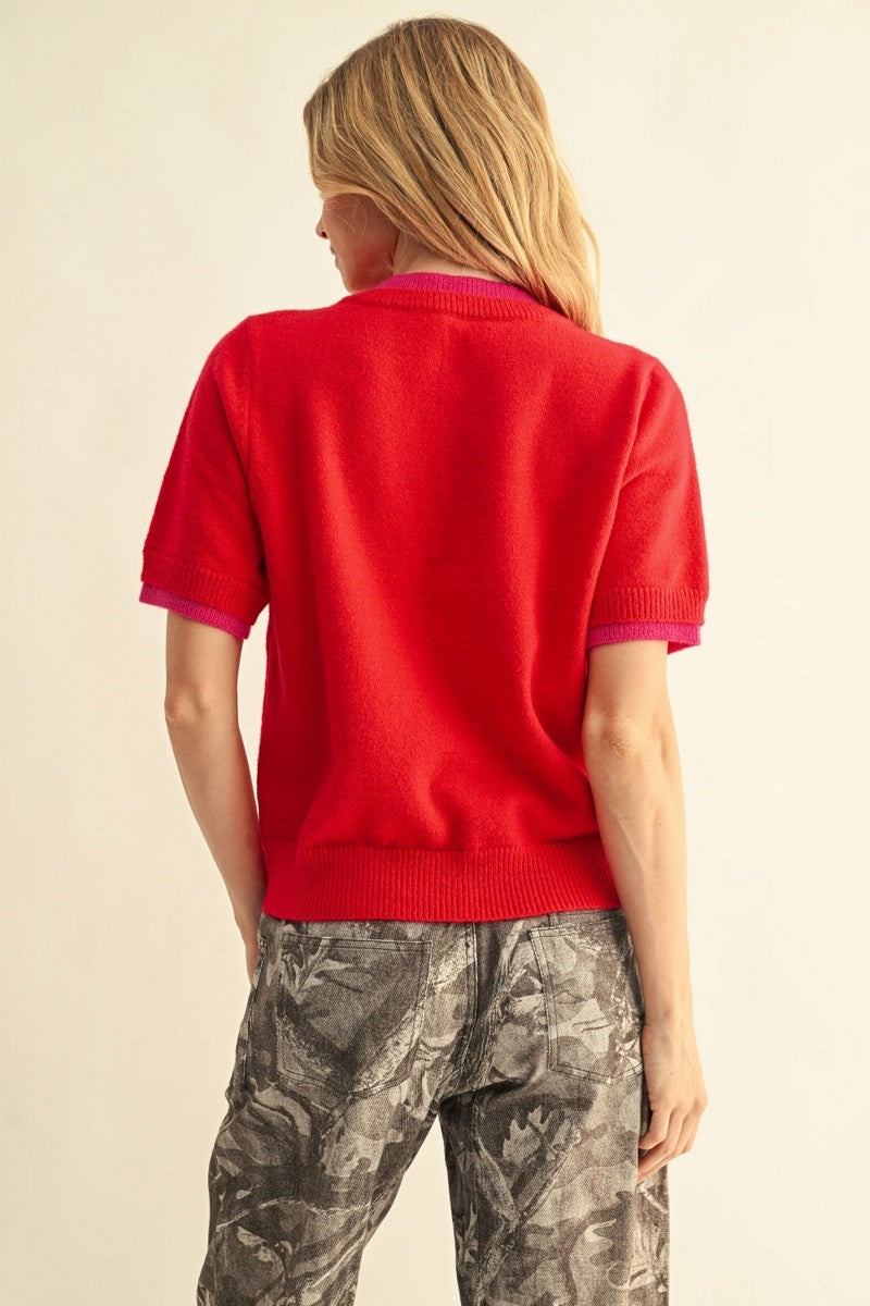 A woman shown from the back wearing a bright red short-sleeve knit top with contrasting magenta trim and gray camouflage pants, against a plain light beige background.